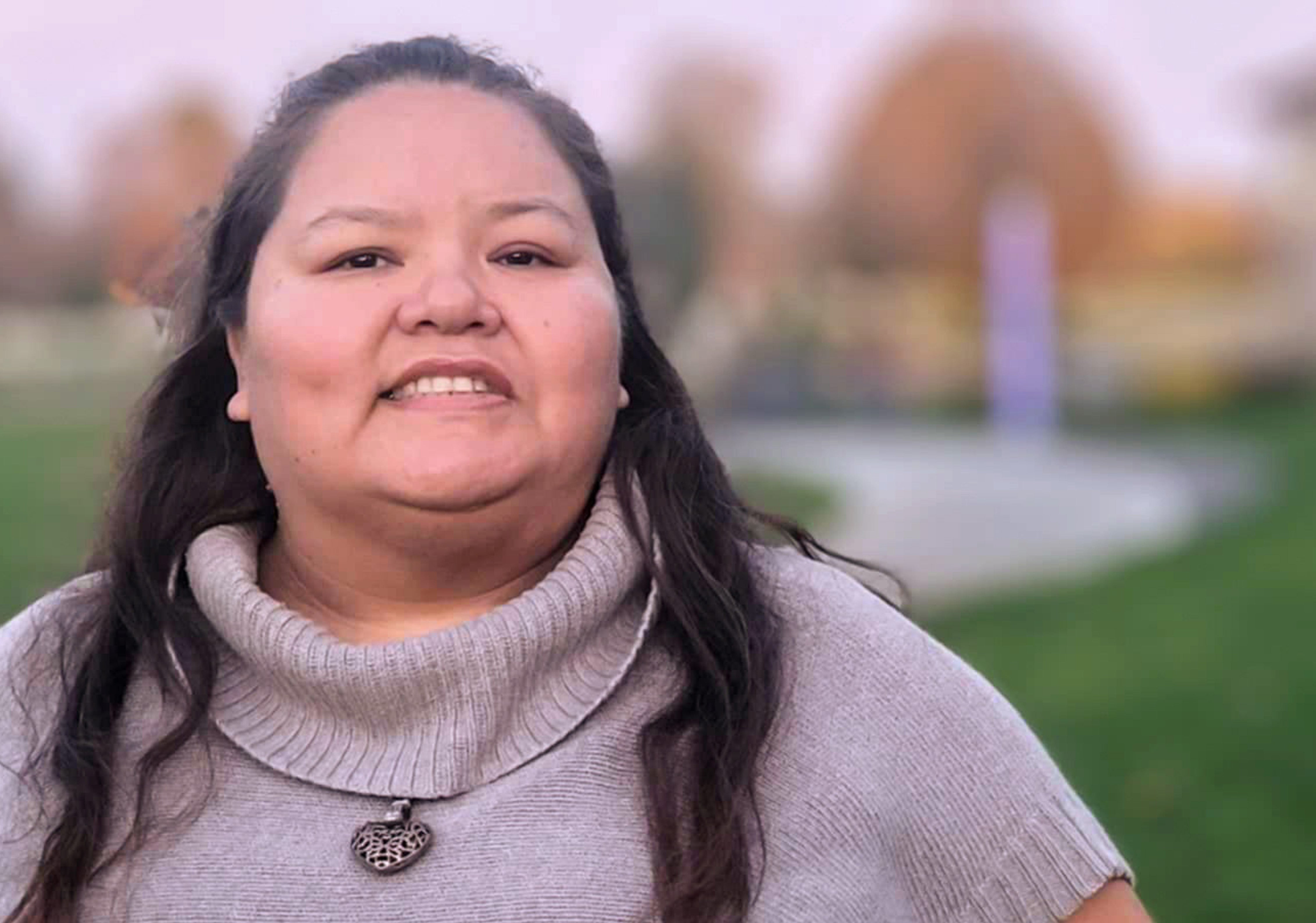 An outdoor portrait photo of a smiling person with long dark hair, wearing a purple short sleeve cashmere sweater and a heart-shaped locket on their chest.